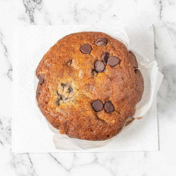 Top-down view of a golden-brown chocolate chip muffin on a white napkin, on a marble surface.