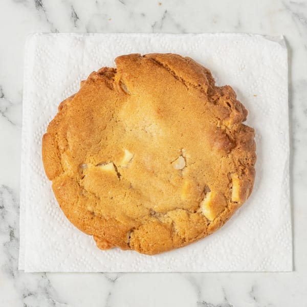 A golden-brown white chocolate chip cookie rests on a white napkin against a marble background.