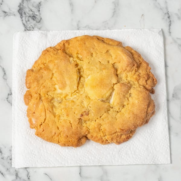 A large, golden-brown cookie with white chocolate chips rests on a white napkin on a marble surface.